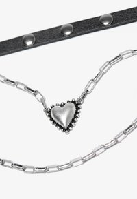Hõbedane südame ripats, mille servad on helmesed, metallkett ja must nahkrihm, mis on kaunistatud metallist nupuhi.