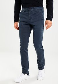 Mörkblå bomullschinos med slim fit, med knäppning, sidofickor och subtil textur. Bärs med vita sneakers.