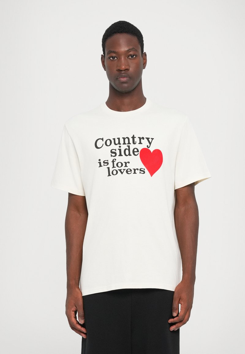 Witte katoenen T-shirt met de tekst "Landelijke omgeving is voor liefhebbers" in het zwart, met een rode hartaccent. Korte mouwen, ronde hals.