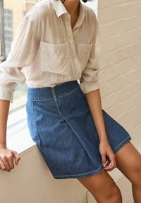 Vaaleanbeige napituspaita, jossa on kaksi etutaskua, yhdistettynä tummansiniseen A-linjaisiin hameeseen, jossa on etukäytävä ja rispaantunut helma.