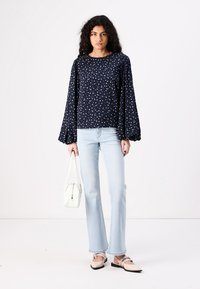 Blusa blu navy con motivo floreale bianco e maniche a palloncino, abbinata a jeans a zampa d'elefante azzurri e ballerine beige. Borsa bianca in mano.
