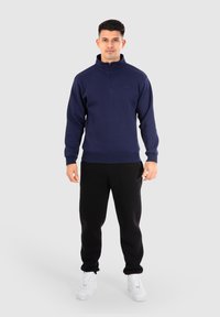 Maglione in pile blu navy con mezza zip e polsini a costine, abbinato a pantaloni tuta neri e sneakers bianche, con un dettaglio logo discreto.