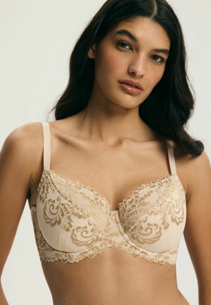 Kobieta w beżowym biustonoszu z miseczkami na fiszbinach, zdobionym kwiatowymi wzorami i regulowanymi ramiączkami, na jednolitym jasnym tle.