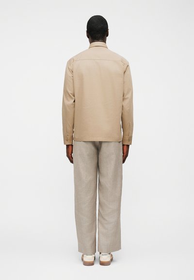 Άνδρας που στέκεται με την πλάτη γυρισμένη, φορώντας μια μπεζ μπλούζα με μακριά μανίκια, ανοιχτό γκρι παντελόνι και λευκά αθλητικά παπούτσια με κείμενο στη φτέρνα.