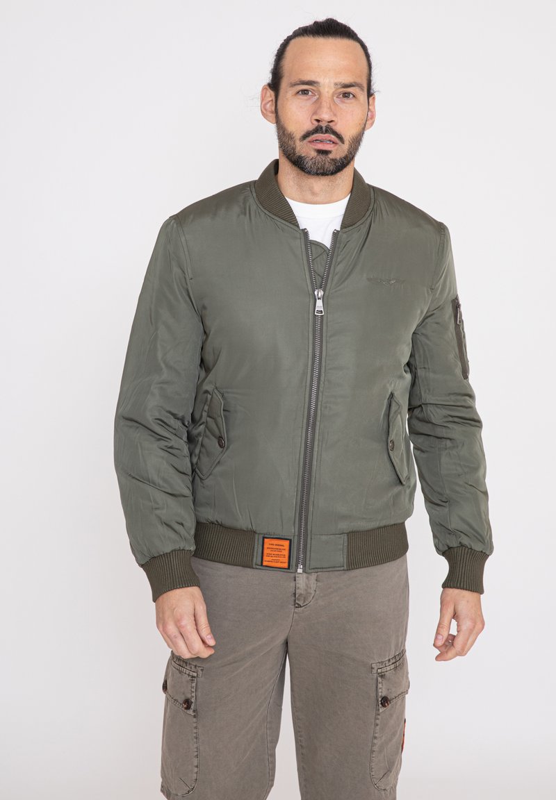 Hombre con barba que lleva una chaqueta bomber verde y pantalones cargo marrones, de pie frente a un fondo liso.