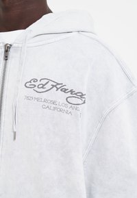 Lys grå hættebluse med lynlås, frontlomme og trykt tekstdesign i mørkere grå. Blødt stof med en struktureret overflade.