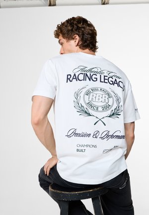 Mann mit lockigem Haar trägt ein weißes T-Shirt mit dem Text und Logo "Racing Legacy", sitzt auf einem schwarzen Hocker und ist von der Kamera abgewandt.