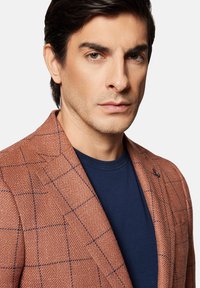 Brązowa marynarka o fakturowanej strukturze w niebieską kratkę, zestawiona z granatowym t-shirtem z okrągłym dekoltem. Marynarka ma klasyczny kołnierz i zapięcie na jeden guzik.