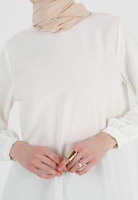 Weiße Bluse mit langen Ärmeln, hohem Kragen und geknöpften Manschetten; kombiniert mit einem beigen Schal. Hände tragen einen goldenen Ring und eine große metallische Manschette.