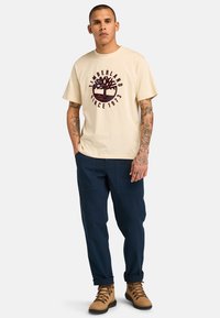 béžové bavlněné tričko s marónovým logem stromu a nápisem "Timberland Since 1973". V kombinaci s námořnickými cargo kalhotami a hnědými turistickými botami.