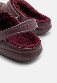 Crocs bourgogne avec doublure pelucheuse, dotés d'une large lanière et d'une semelle texturée. Le design intègre des trous de ventilation et des matériaux doux.
