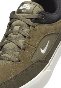 Nike SB Malor kedas ar olīvzaļas zamšas un auduma augšdaļu, baltu Swoosh logo, teksturētu šņorēšanas daļu un balto gumijas zoli.