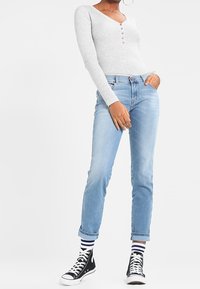 Maglietta henley grigia a maniche lunghe con bottoni, jeans di denim blu chiaro e sneakers nere alte con calzini a righe, in piedi su uno sfondo neutro.
