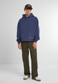 Marineblauwe hoodie met een kangaroo-zak aan de voorkant, gecombineerd met olijfgroene broek en grijze sneakers. De pet is zwart met een logo.