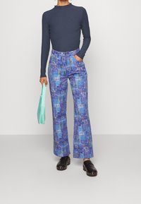 Pantalon évasé à motifs bleus avec divers graphismes, associé à un haut à manches longues côtelé, et un sac à main vert menthe. Des sabots noirs complètent le look.