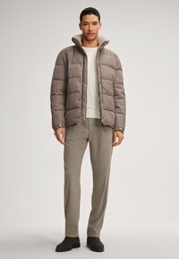 Pufferjack in gedempt bruin, met een gewatteerd ontwerp en een zachte grijze capuchon, gedragen over een licht sweatershirt en beige broeken. Bruine laarzen maken de look compleet.