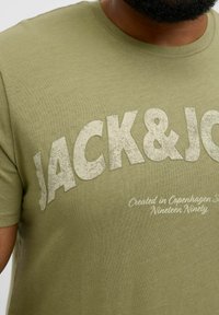 Olivgrünes Baumwoll-T-Shirt mit strukturiertem weißen Schriftzug "JACK&JOE" und kleinerem Text darunter. Kurze Ärmel und runder Halsausschnitt, glattes Gewebe.