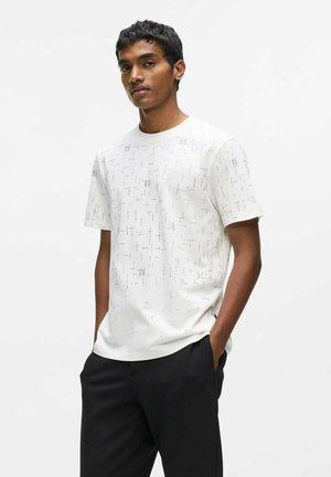 Jonge man met donker haar die een wit, kort mouw T-shirt met patroon en zwarte broek draagt, staat met zijn handen in de zakken tegen een witte achtergrond.