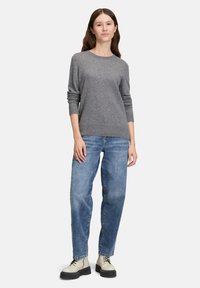 Junge Frau steht, trägt einen grauen Langarmpullover, lockere blaue Jeans und beigen Schnürboots vor einem einfarbigen hellen Hintergrund.