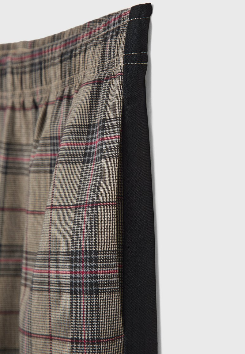 Pantaloni a quadri con un motivo beige, nero e rosso, dotati di una vita elasticizzata e pannelli laterali in tessuto nero. Texture liscia.