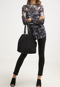 Schwarze Bluse mit Blumenmuster und langen Ärmeln, kombiniert mit schwarzen Skinny-Jeans und einer schwarzen Wildleder-Shoppertasche. Offene schwarze Mules runden das Outfit ab.