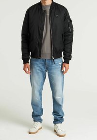 Man in een zwarte bomberjack over een grijze sweater, lichtblauwe distressed jeans en witte sneakers met bruine zolen.