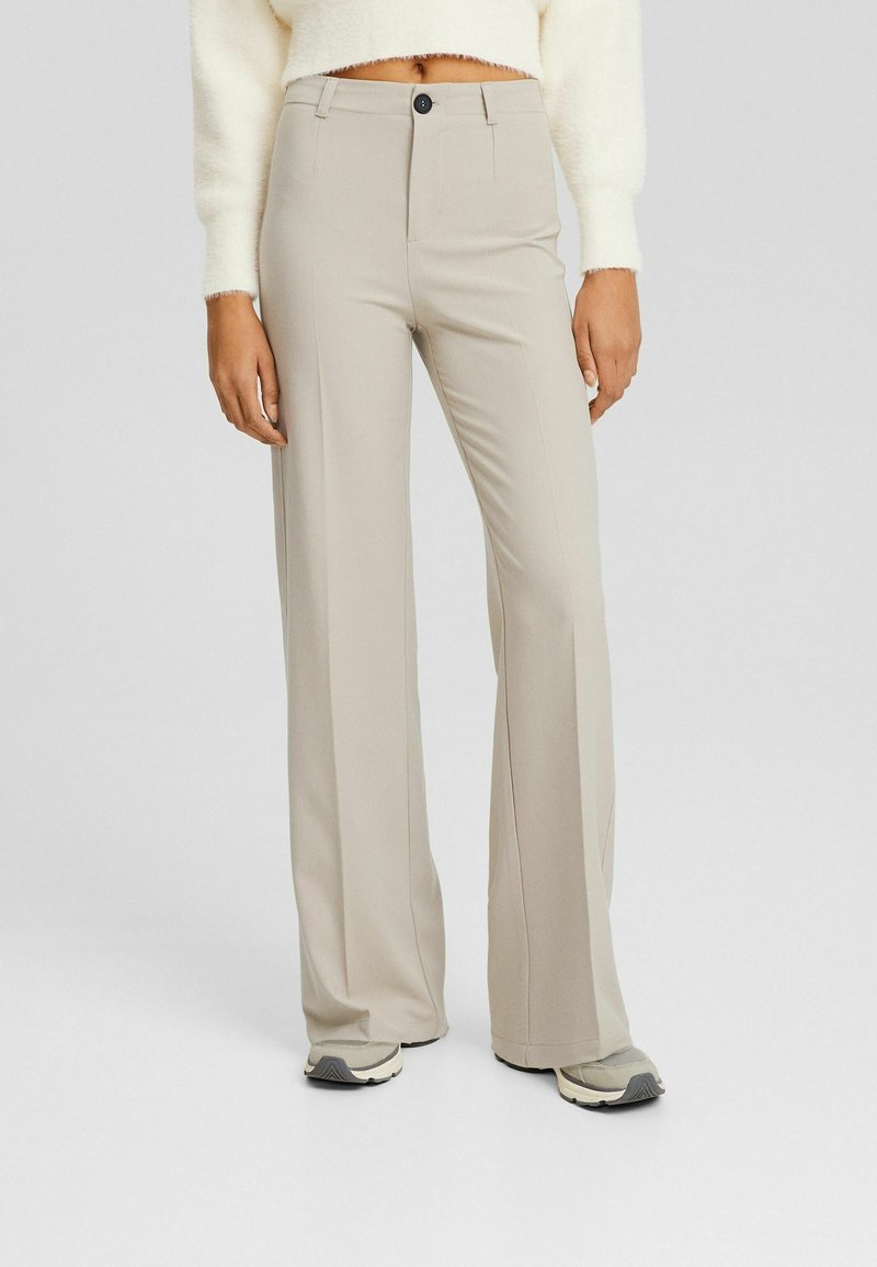 Pantalon beige à jambes larges avec bouton et fermeture éclair devant, porté avec un pull court blanc et des baskets grises sur un fond uni.