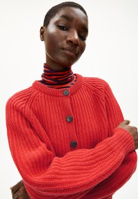 Rode gebreide cardigan met ribbelstructuur, voorzien van zwarte knopen, ronde halslijn en schouders die zijn laten zakken, gedragen over een rood en blauw gestreepte coltrui.