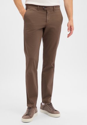 Man wearing brown slim-fit pants and taupe sneakers with white soles, standing with one hand in pocket against plain background.