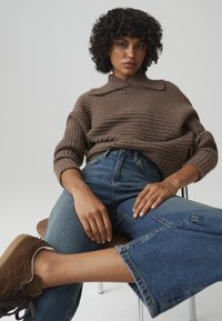 Brown ribbed sweater with a collar, paired with light blue wide-leg jeans. Model wears brown suede sneakers, seated on a wooden stool.