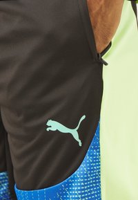 Schwarze Sportshorts mit grünen Akzenten, die einen blauen gemusterten Abschnitt und ein hellblaues Puma-Logo auf dem linken Bein aufweisen.