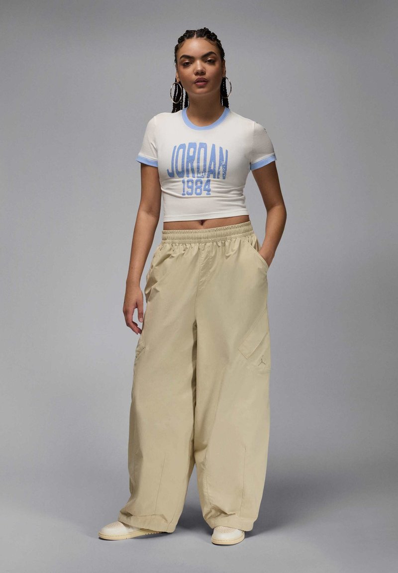 T-shirt blanche coupée avec bordures bleues et graphique "JORDAN 1984", associée à un pantalon cargo beige ample avec poches latérales et taille élastique.