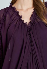 Fioletowa bluzka z dekoltem w kształcie litery V, falbaniastymi krawędziami i plisowanym przodem. Wykonana z lekkiego, przezroczystego materiału o miękkiej teksturze.