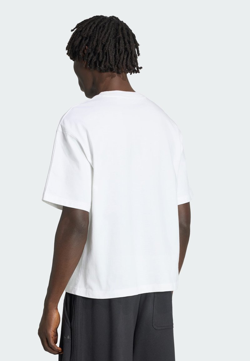 Uomo con dreadlocks corti che indossa una semplice t-shirt bianca a maniche corte e pantaloncini neri, di spalle su uno sfondo chiaro.