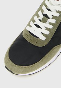 Sneaker met olijfgroen suède, zwarte stoffen bovenkant, witte veters en witte zool, getoond vanuit een boven-frontal close-up perspectief.