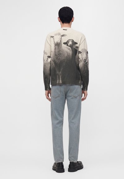 Homme portant un pull gris avec un motif de mouton noir et blanc dans le dos, jean bleu clair et chaussures noires, debout de dos.