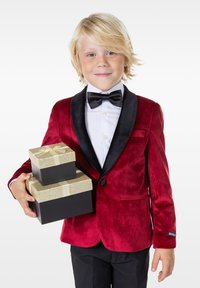 Chaqueta de esmoquin de terciopelo rojo con solapas negras, camisa blanca y pajarita negra. Sosteniendo dos cajas de regalo, una negra y una dorada con cinta.
