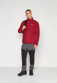 Veste en polaire rouge avec fermeture éclair intégrale, pantalon de randonnée noir et gris, et chaussures de randonnée noires. Le modèle tient un sac noir, se tenant sur un fond uni.