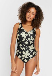 Bañador negro con estampado floral y diseño envolvente; presenta patrones de flores verde claro, tirantes laterales ajustables y un escote en V. Tejido suave y elástico.