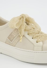 Sneaker beige avec de grosses lacets en mailles métalliques dorées noués en nœud et un détail pailleté sur le côté.