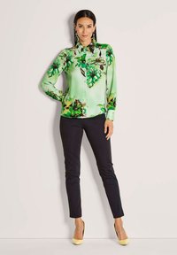 Vrouw in een groene bloempatronen blouse met knopen, zwarte slim fit pants, gele hakken en grote hangende oorbellen, staand tegen een witte muur.