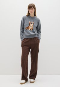 Grijze gebreide trui met een kattenontwerp, gecombineerd met bruine geruite broeken. Model draagt witte sneakers. Eenvoudige, relaxte pasvorm.