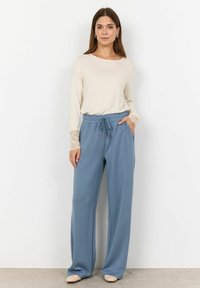 Maglietta beige a maniche lunghe con pizzi ai polsini abbinata a pantaloni ampi azzurri con vita regolabile e tasche laterali.