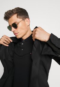 Crna jakna nosena preko crnog polo majice s dugmadi. Model nosi sunčane naočale s zelenim nijansama. Glatka tekstura, strukturirani dizajn.