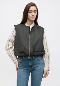 Šedá prešívaná vesta na zips vpredu s dvoma prednými vreckami. Nosená cez bielu košeľu s vertikálnymi pruhmi, doplnená modrými džínsami.