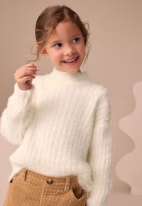 Pull blanc en maille côtelée avec un col montant côtelé et une texture douce, associé à un pantalon en velours côtelé couleur beige avec un détail de bouton rond.