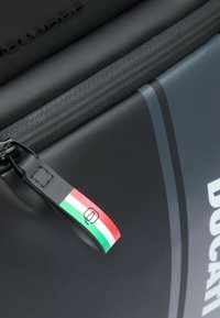 Bolsa negra con cremallera y superficie texturizada. Presenta una etiqueta de tela con franjas rojas, blancas y verdes, y un logotipo en relieve en el borde.