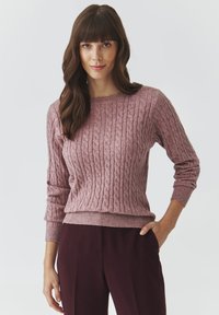 Cable-knit sweater in soft pink with round neckline, ribbed cuffs, and hem. Worn with dark burgundy pants. Texture is soft and cozy.
