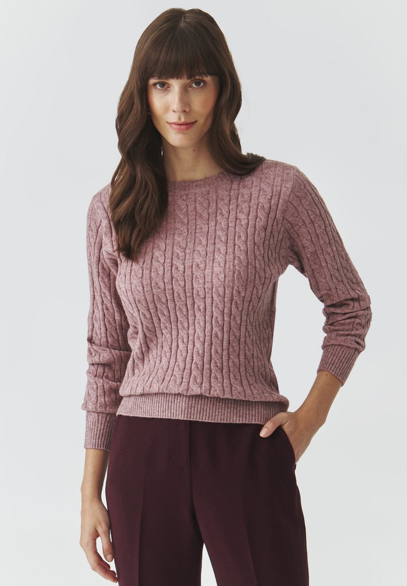 Cable-knit sweater in soft pink with round neckline, ribbed cuffs, and hem. Worn with dark burgundy pants. Texture is soft and cozy.