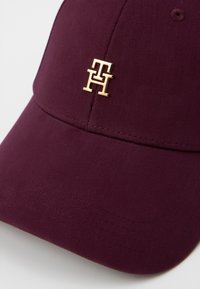 Burgundijas krāsas cepure, izgatavota no auduma, ar zelta logotipa akcentu. Dizains ietver izliekto malu un teksturētu materiālu.